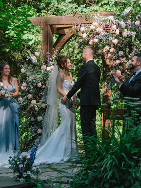An artistic portrait shot through an open wooden doorway, capturing a bride holding her bouquet in a rustic sunlit bedroom.
