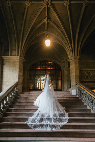 Dramatic back view of a bride with a long lace cathedral-length wedding veil cascading down the monumental stone stairs beneath the vaulted ceilings at Knox College.