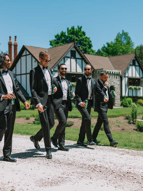 Joyful candid shot of a groom and his groomsmen in black tuxedos walking down the gravel driveway with the historic Tudor estate manor in the background on a sunny day.