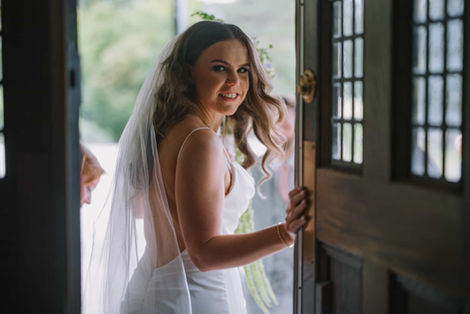 Bride in a white lace wedding dress smiling while standing by the grand wooden front door of the historic estate.
