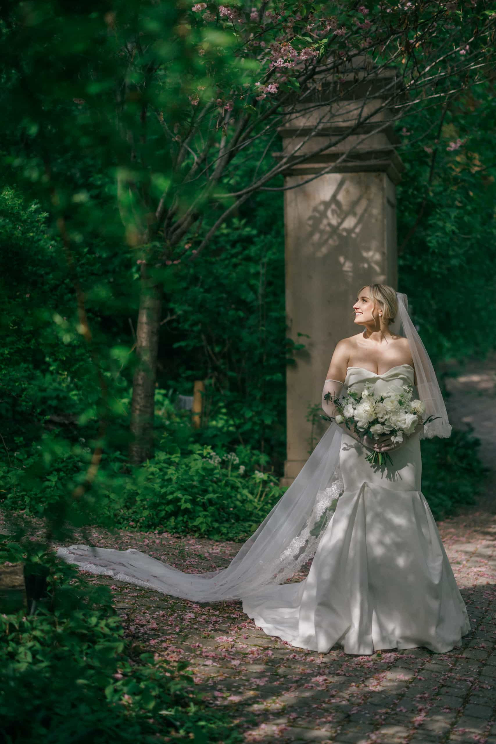 A bride with a long flowing veil walking down a lush green garden pathway holding a bouquet, captured by a Toronto wedding photographer.
