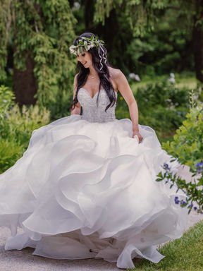 A bride wearing a dramatic ruffled ballgown and a green flower crown walking along a stone pathway in a lush garden.