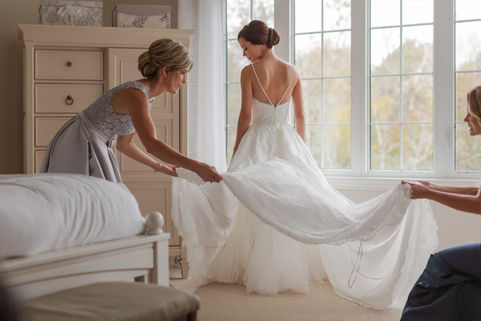 A candid documentary moment showing a bridesmaid carefully arranging the bride's tulle wedding dress train in a sunlit bedroom.