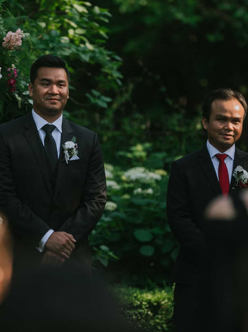 A documentary photograph capturing a groom smiling emotionally at the altar while watching his bride walk down the outdoor garden aisle.