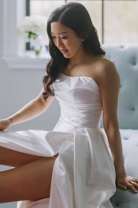 A beautifully lit indoor bridal portrait of a smiling bride sitting by a bright window in a modern gown.
