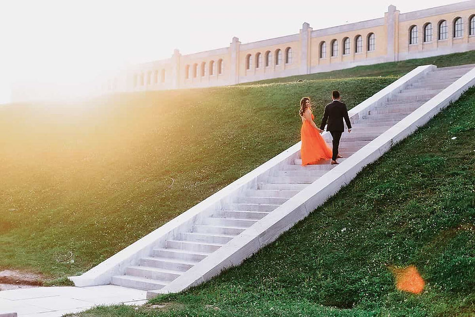 A couple in formal attire walks up sunlit stairs by a grassy hill, with an orange dress and a distant building in the background. Romantic mood.