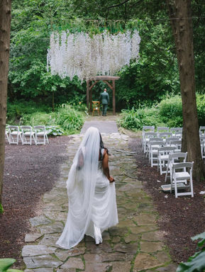 A stunning portrait of a bride standing elegantly near a massive wooden bed frame with natural window light illuminating her face.