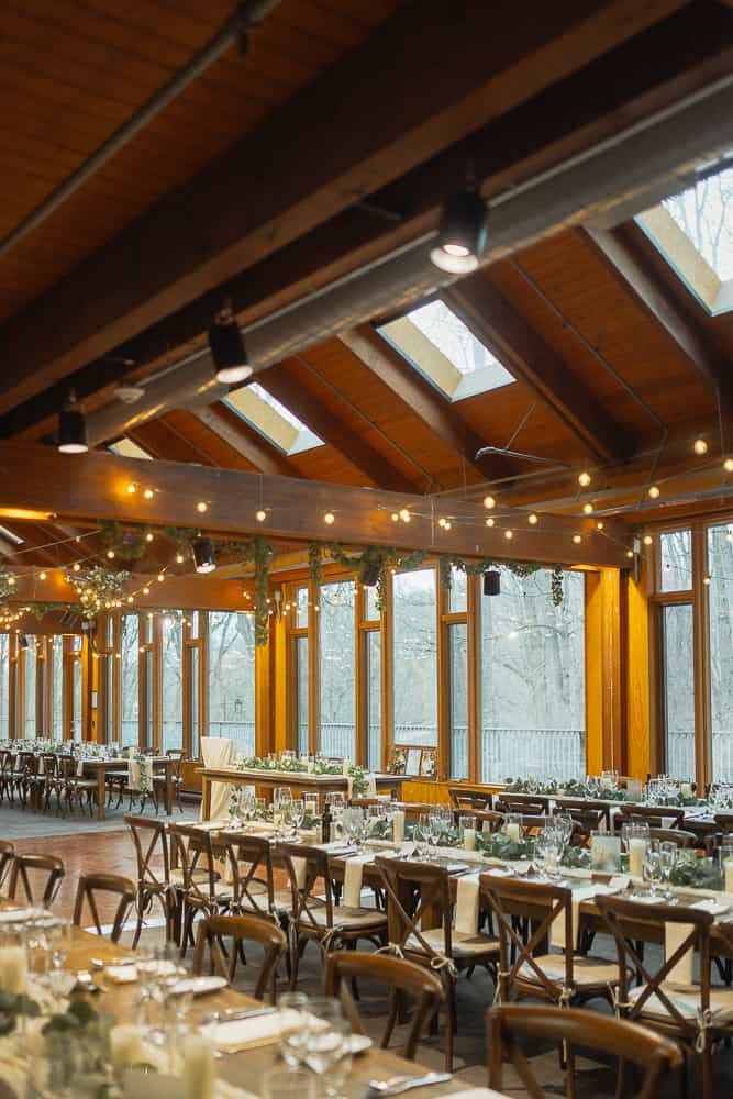Elegant dining hall with wooden beams, skylights, and string lights. Tables set with white linens and glassware, creating a warm, inviting mood.