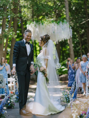 A beautiful outdoor wedding ceremony in progress deep within a secluded forest clearing surrounded by tall pine trees and seated guests.