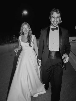 lack and white shot of a happy bride and groom walking outside at night, holding drinks and smiling during their reception.
