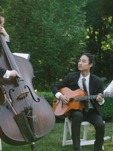 A live musical trio consisting of a double bass player, guitarist, and violinist performing outdoor ceremony music on the estate lawns.