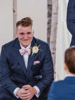 A candid documentary moment of a groom in a navy suit smiling warmly as a young ring bearer approaches the wedding altar.