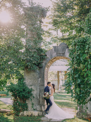 A wedding couple embracing beneath a historic, vine-covered stone arch monument on the expansive heritage grounds of the Guild Inn Estate.