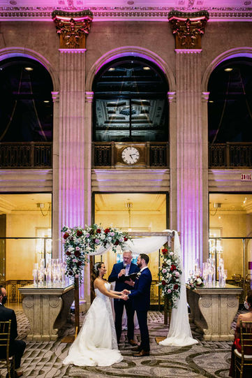 A beautiful wedding altar setup framed by towering historic arches with purple uplighting inside the Grand Banking Hall.