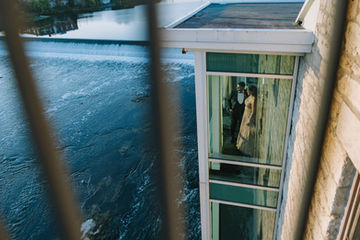 A highly artistic wedding photograph capturing the reflection of a bride and groom in a glass window, juxtaposed creatively with rushing blue water in the foreground.

