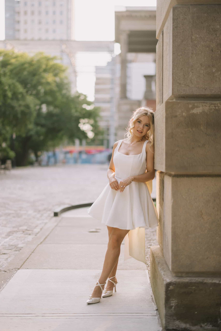  A stylish woman in a short white dress smiles as she poses against the historic architecture of Osgoode Hall for her engagement portrait.