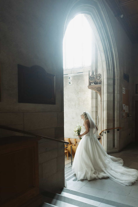 Romantic bridal portrait of a bride in a white ballgown standing near a sun-drenched arched doorway within the stone walls of Trinity College Chapel.