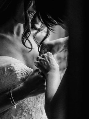 A close-up black and white candid shot of hands carefully buttoning the delicate lace back of a bride's off-the-shoulder wedding dress.
