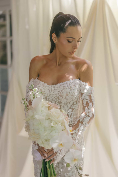 Close-up bridal portrait highlighting an intricate off-the-shoulder lace wedding dress and a delicate white floral bouquet.