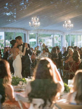A newlywed couple shares their first dance surrounded by seated guests in a beautiful, sunlit glass-walled reception tent.