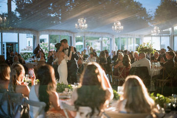 A newlywed couple shares their first dance surrounded by seated guests in a beautiful, sunlit glass-walled reception tent.