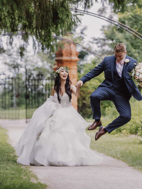 A hilarious and creative candid shot of a groom doing a mid-air jump next to his laughing bride on a garden path.