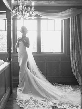 Elegant black and white bridal portrait of a bride standing by a large paned window beneath a luxury crystal chandelier, showcasing her long lace train on the hardwood floor at Erin Estate.