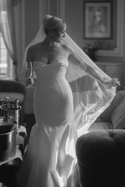 Black and white photograph of a bride from behind, showcasing her fitted wedding dress and holding a flowing veil in an elegant room.