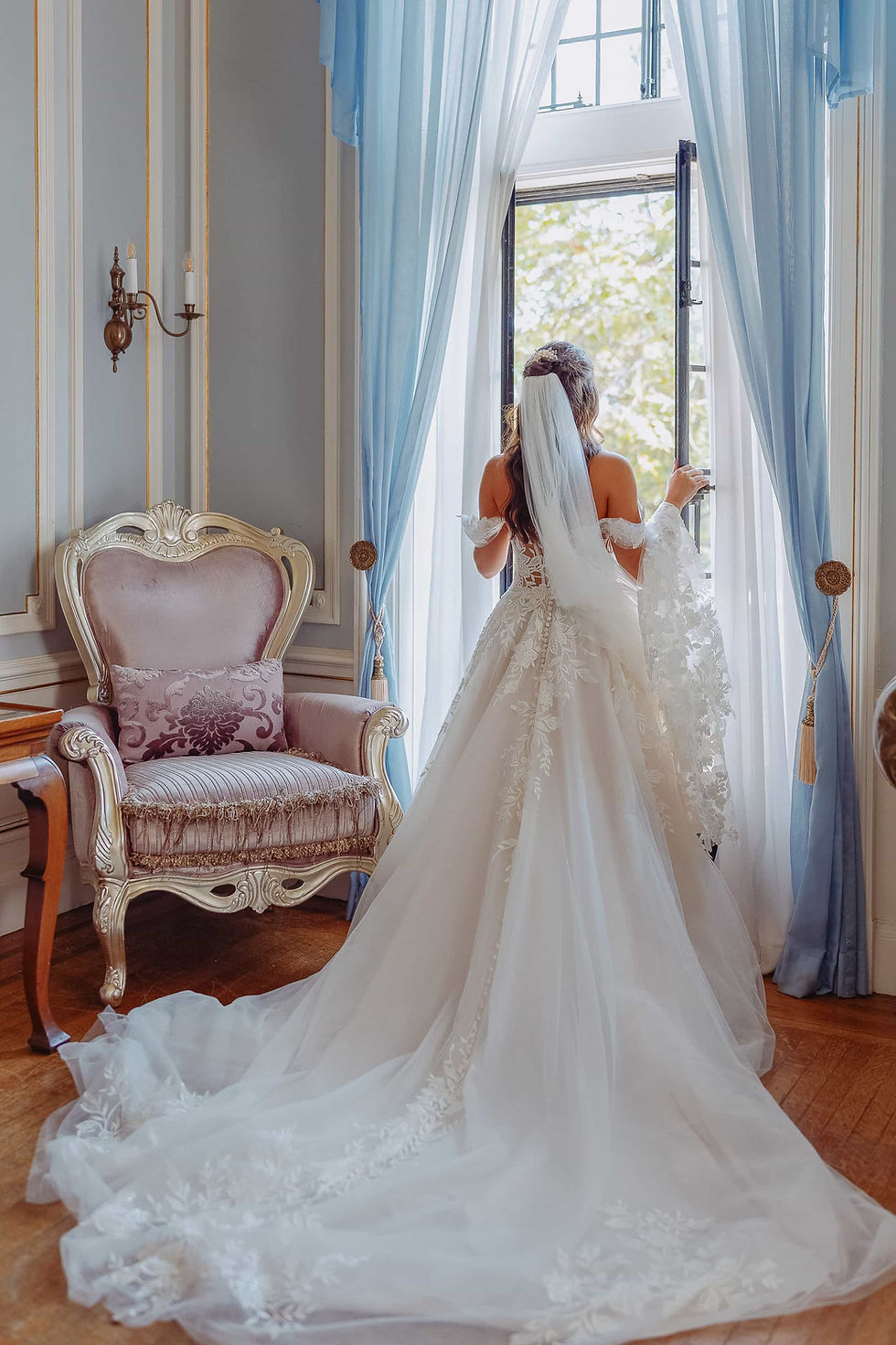 A fine-art bridal portrait focusing on the intricate lace details of a wedding gown. The bride stands in soft window light next to an ornate pink chair in a luxurious Toronto bridal suite.