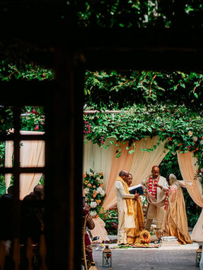 A romantic outdoor wedding ceremony framed creatively through a dark wooden pergola covered in lush green vines.