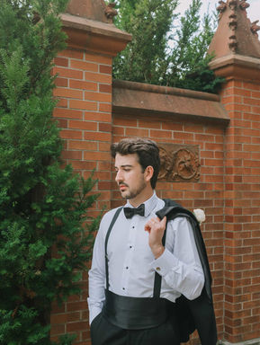 A handsome groom posing with his suit jacket draped casually over his shoulder against a historic red brick pillar wall.