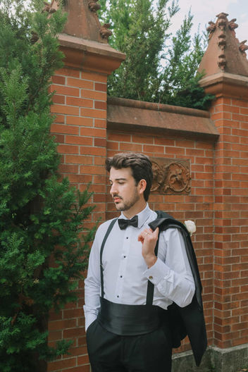 Editorial groom portrait of a groom in a classic tuxedo leaning against a textured historic brick wall at the estate.