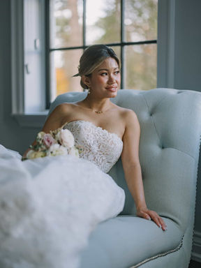 A beautiful indoor portrait of a bride holding her bouquet while sitting relaxed in a plush grey armchair next to a large grid window.