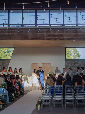 Wide-angle interior photograph of an elegant indoor wedding service inside the contemporary glass-walled pavilion at the Guild Inn Estate, featuring warm wood ceilings and hanging string lights.