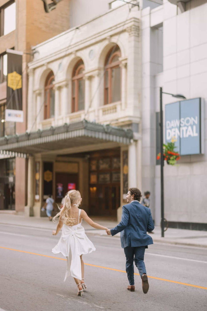 A couple holds hands and runs down a city street in Toronto, away from the camera, with a historic theater in the background.