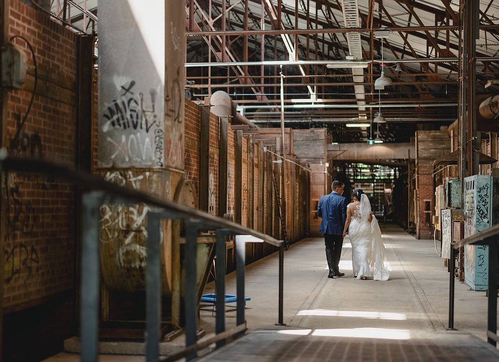 Bride and groom walking in an industrial warehouse with brick walls and graffiti. Sunlight filters through the ceiling, creating a romantic mood.
