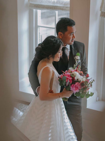 A romantic portrait of a bride and groom standing together on a winding wooden staircase beside a bright, tall estate window.