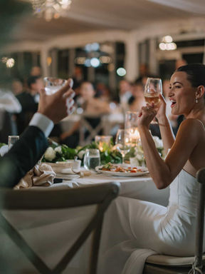A documentary wedding photograph of a bride laughing and raising her glass during a dinner toast inside the marquee tent.