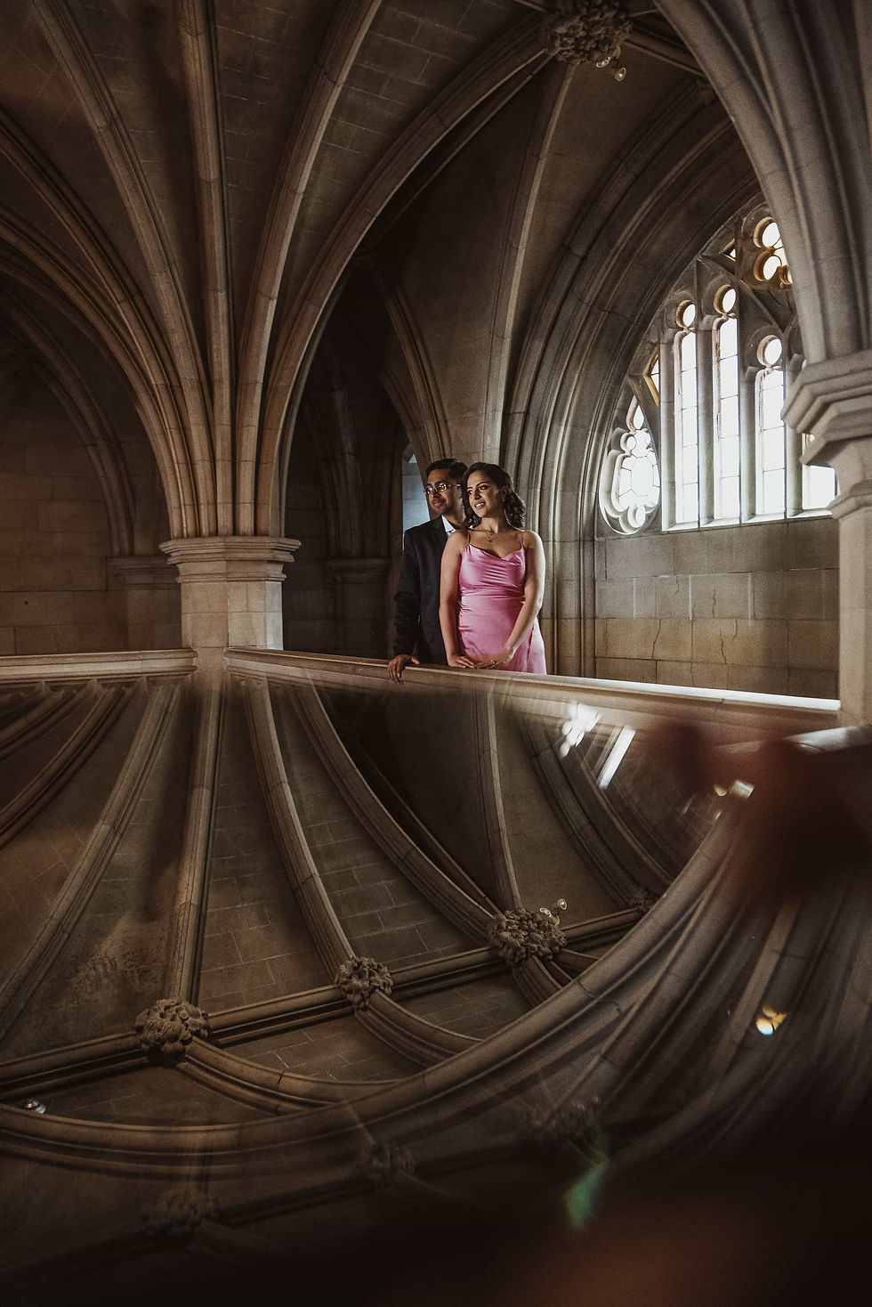 A Toronto wedding photographer captures an elegant couple standing on a stone balcony within a grand Gothic-style interior, featuring dramatic vaulted ceilings and a delicate arched window.