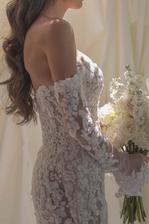 A soft, naturally lit profile portrait of a bride wearing an off-the-shoulder long-sleeve lace gown holding a bridal bouquet.