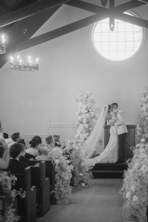 Black and white photograph of a bride and groom kissing at the altar surrounded by tall white floral arrangements.