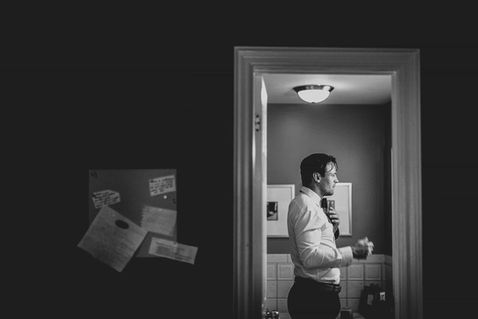 Cinematic black and white candid of a groom adjusting his tie, framed through a doorway for a classic documentary feel.