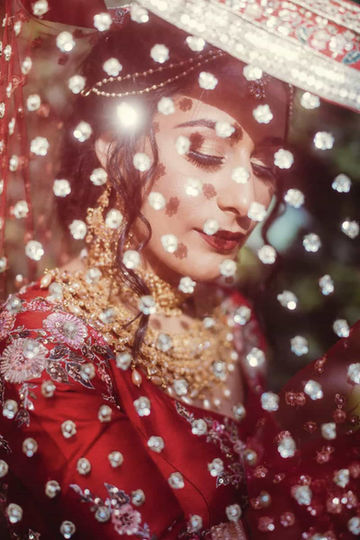 A stunning, artistic close-up portrait of a South Asian bride looking down, photographed creatively through the sparkling, out-of-focus beads of her red veil.