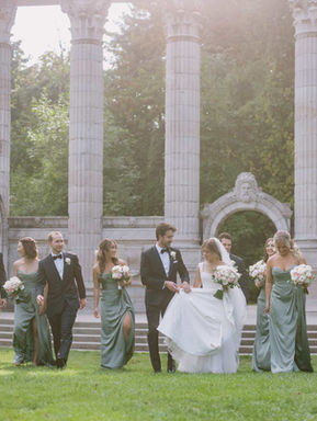 A wide landscape photograph of a full bridal party in sage green dresses and black suits walking past the iconic stone pillars of the Greek Theatre.