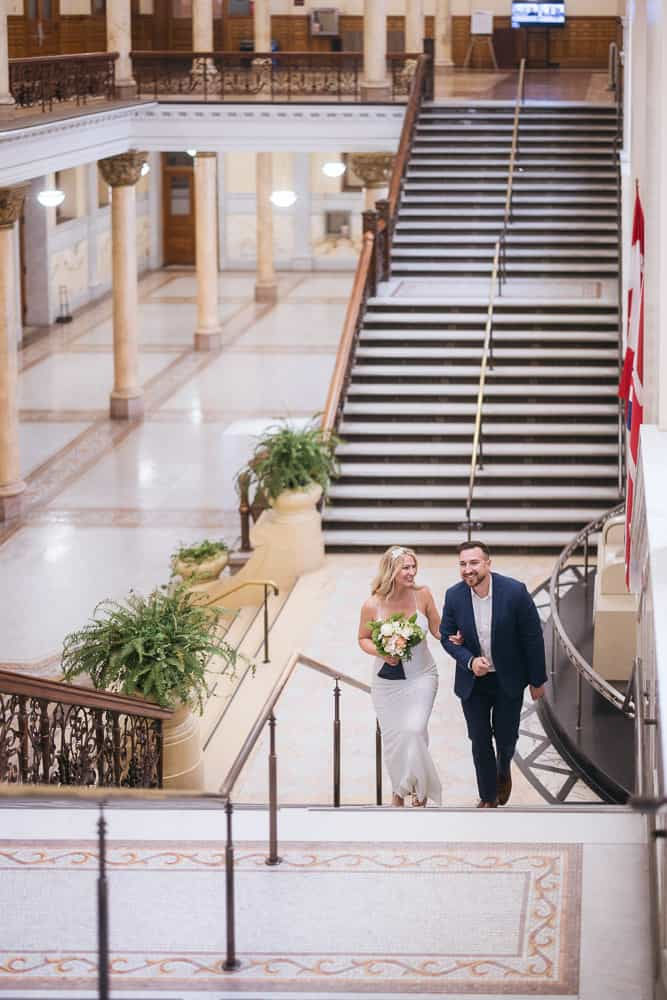 A couple joyfully ascends a grand staircase in an elegant building. The woman holds a bouquet, and plants line the ornate railing.