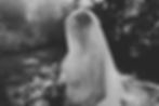 Black-and-white portrait of a bride in lace veil holding a bouquet outdoors, looking down calmly in soft sunlight.