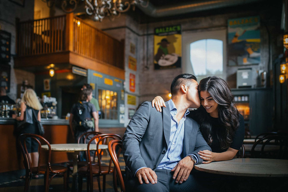 Couple in a cafe, man kissing woman's cheek. Cozy setting with dim lighting and Balzac posters. Warm, intimate mood.