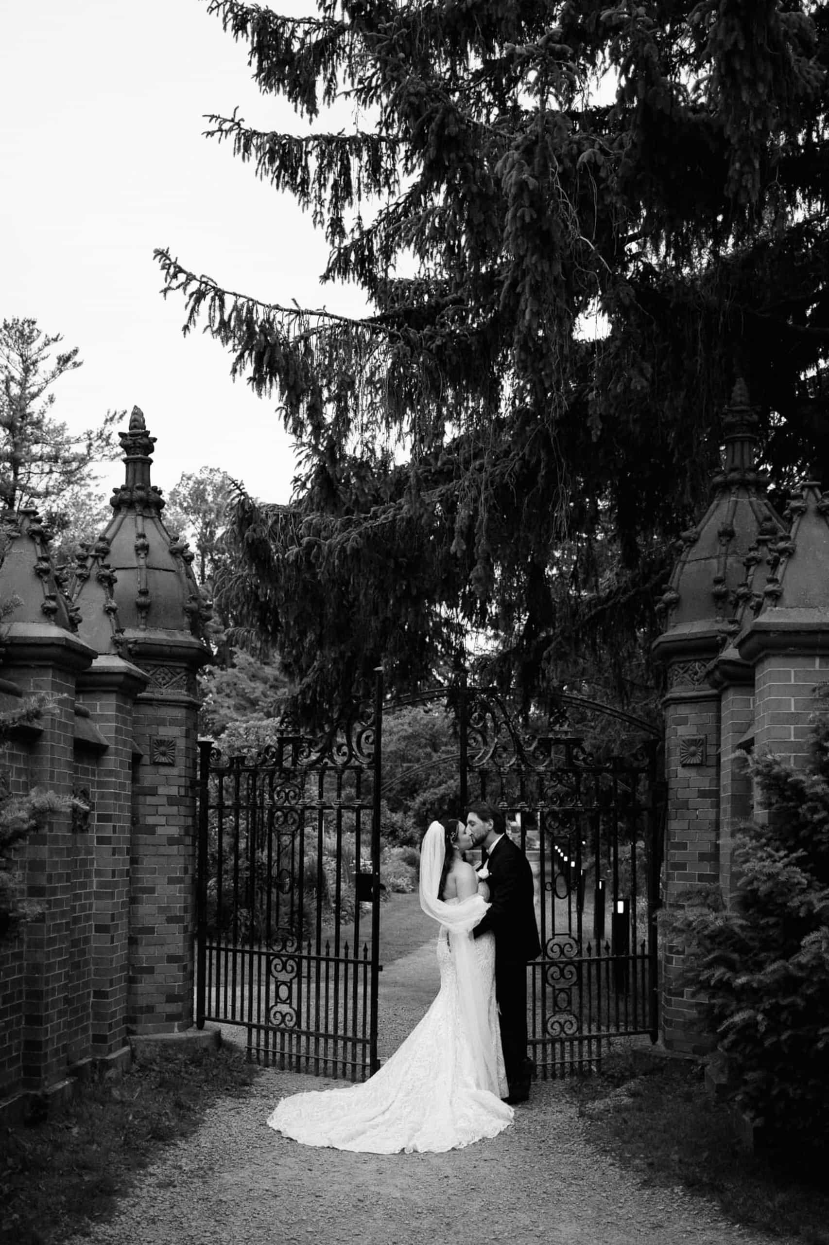 Black and white editorial wedding portrait of a bride and groom standing by the historic stone and wrought-iron gates at Guild Inn Estate in Toronto