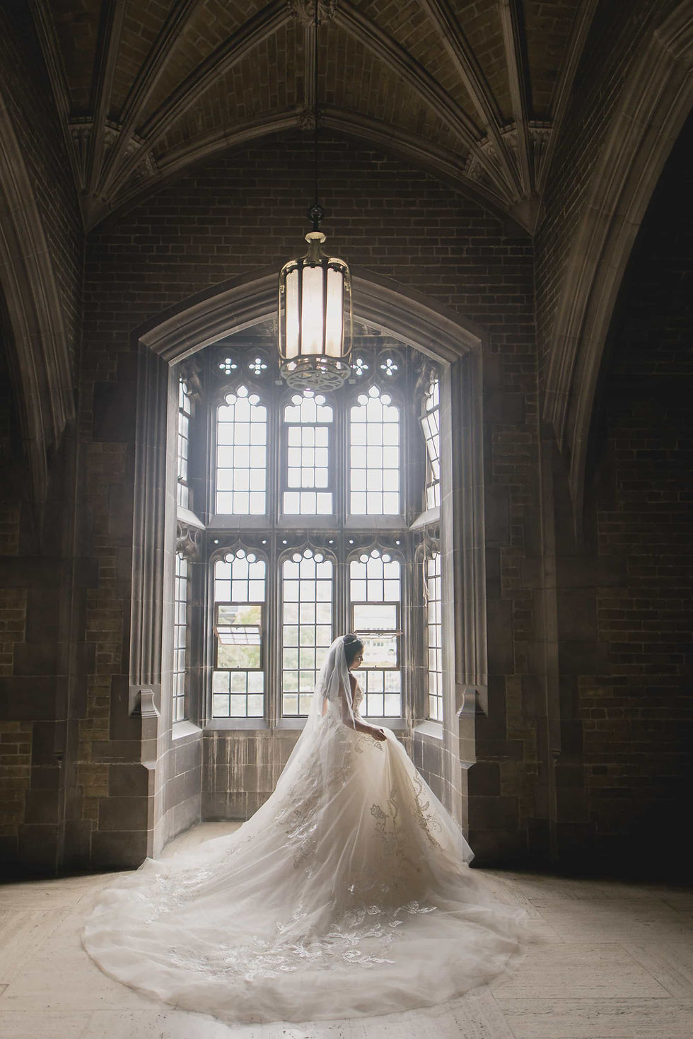 Editorial wedding photography framing the dramatic vertical scale and gothic architecture of the massive stone window inside Hart House Great Hall