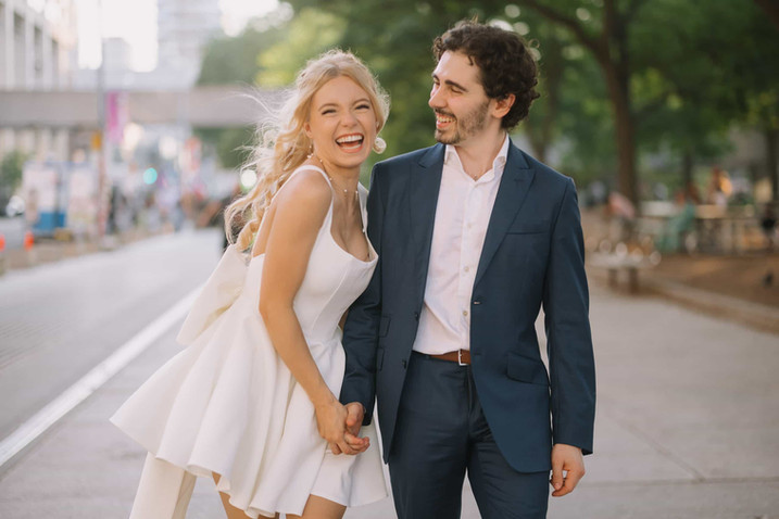 A bride-to-be laughs joyfully while holding her fiancé's hand on a city street during their engagement photoshoot.